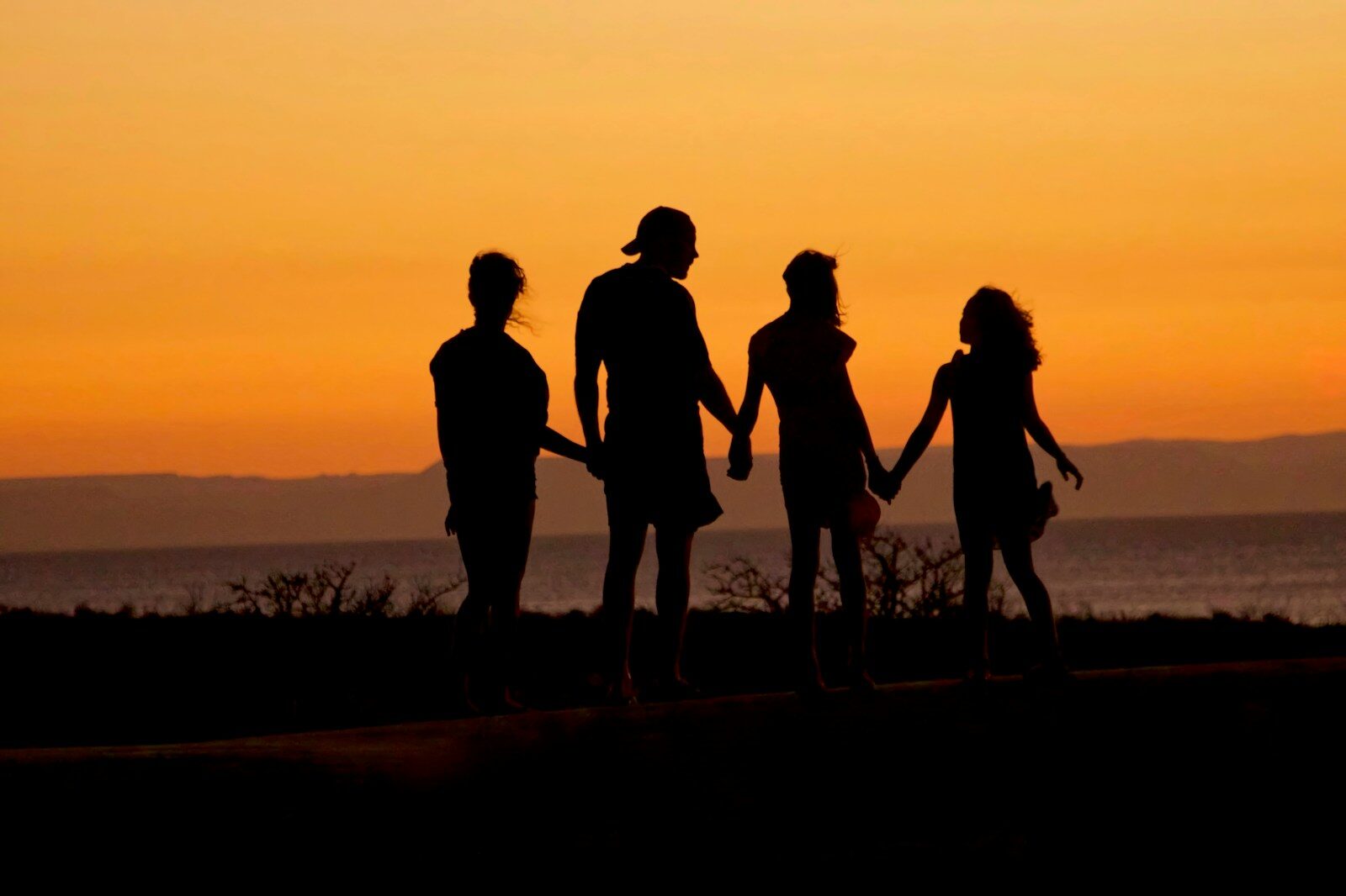 A family in silhouette holding hands against a warm orange sky by the sea